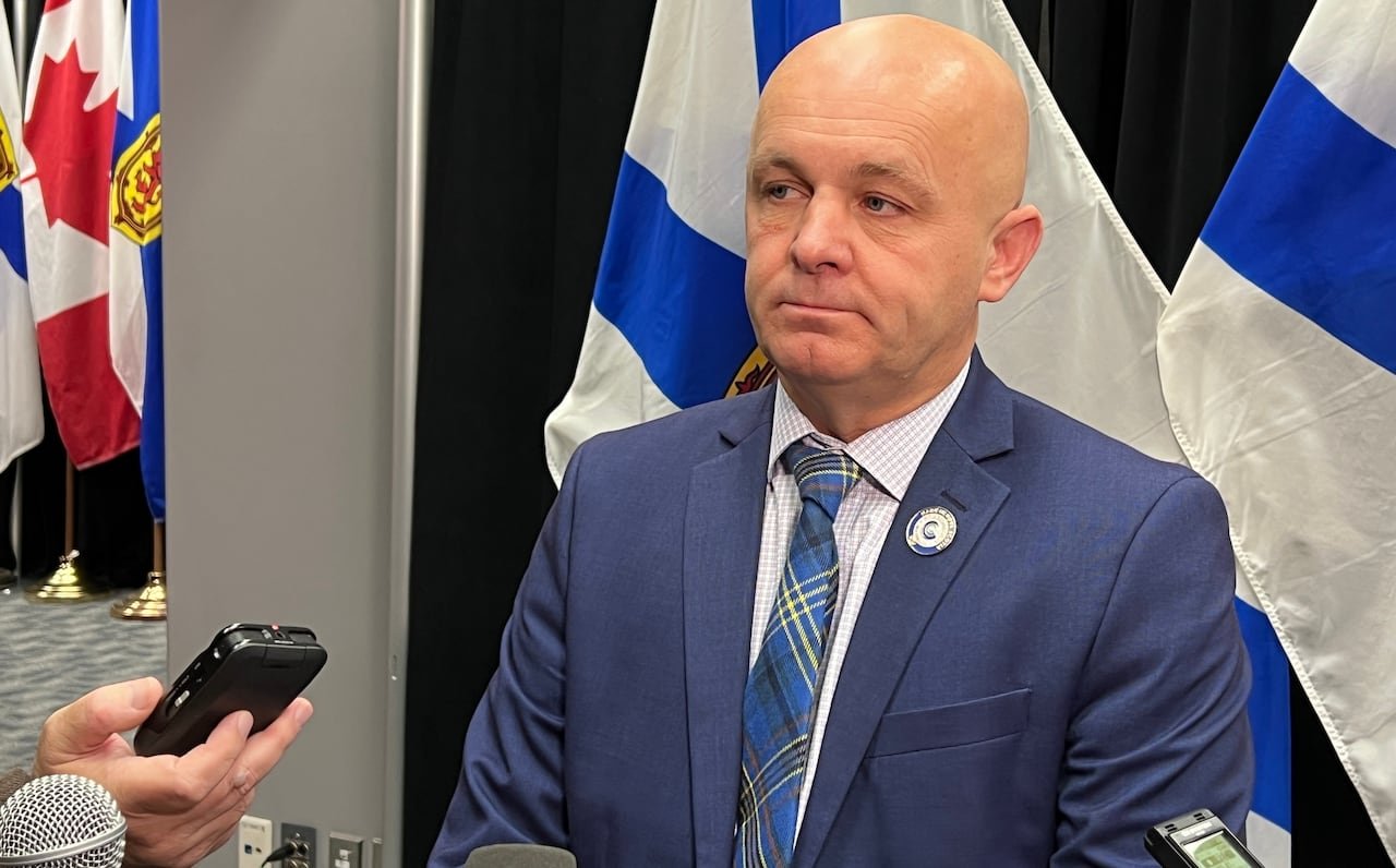default-495 A bald man in a suit and tie stands in front of Nova Scotia flags.