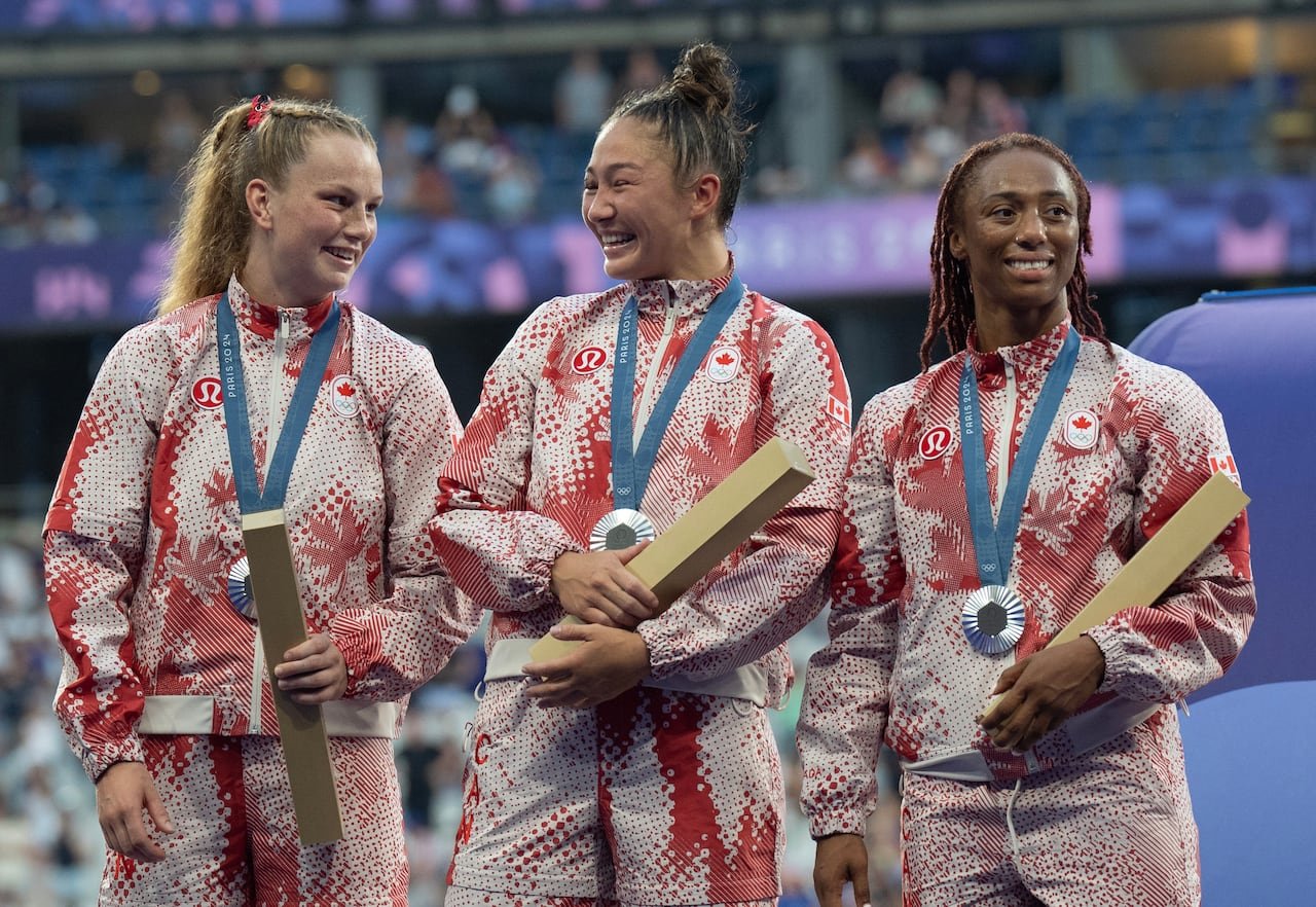 Three women wearing Team Canada jackets smile with silver Olympic medals around their necks.