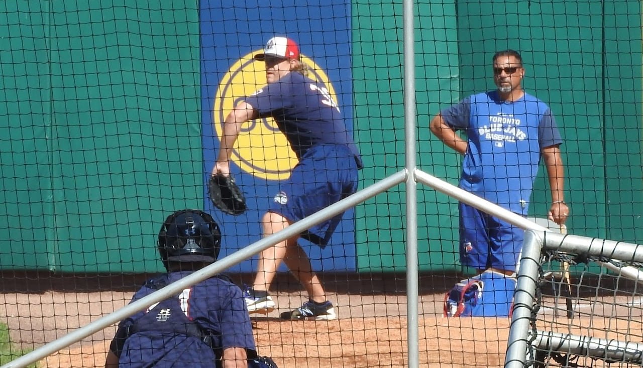 A pitching coach looks on as a player throws to a catcher.