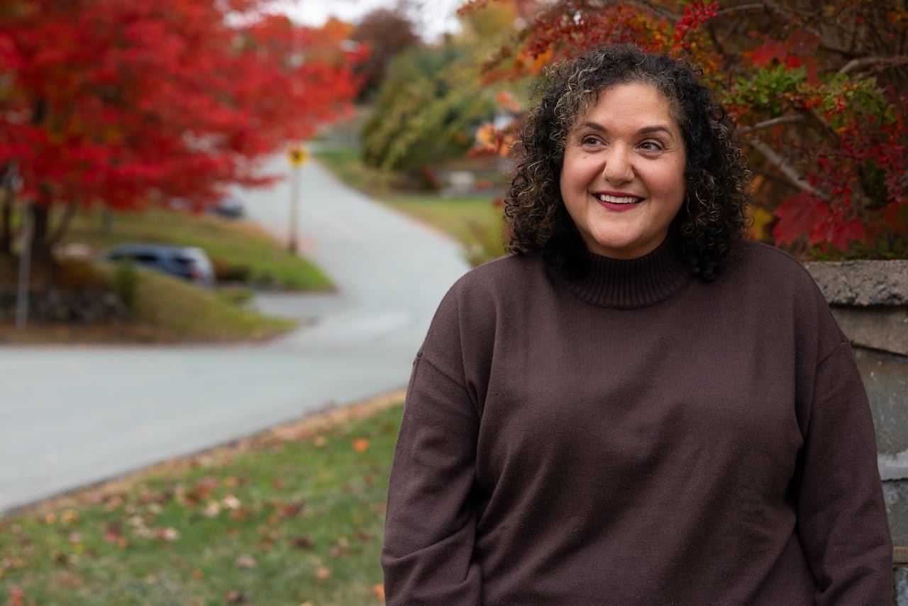 default-430 A woman in a brown sweater sits outdoors with a street sloping up behind her and colourful trees in the distance.