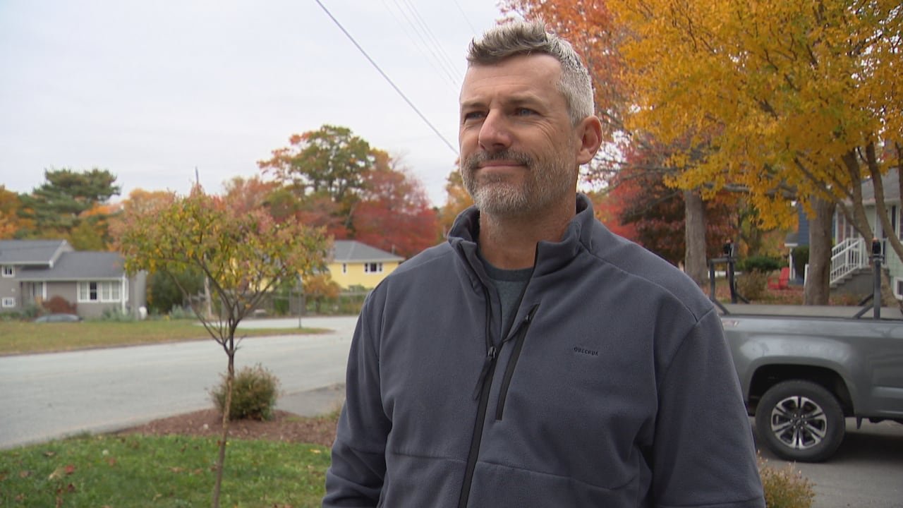 default-431 A man in a grey zip-up sweater stands with houses and trees visible behind him.