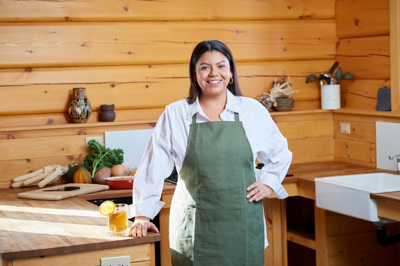 Woman stands at butcher block counter, wearing green apron with a beverage. Log walls in the background make for a rustic kitchen.
