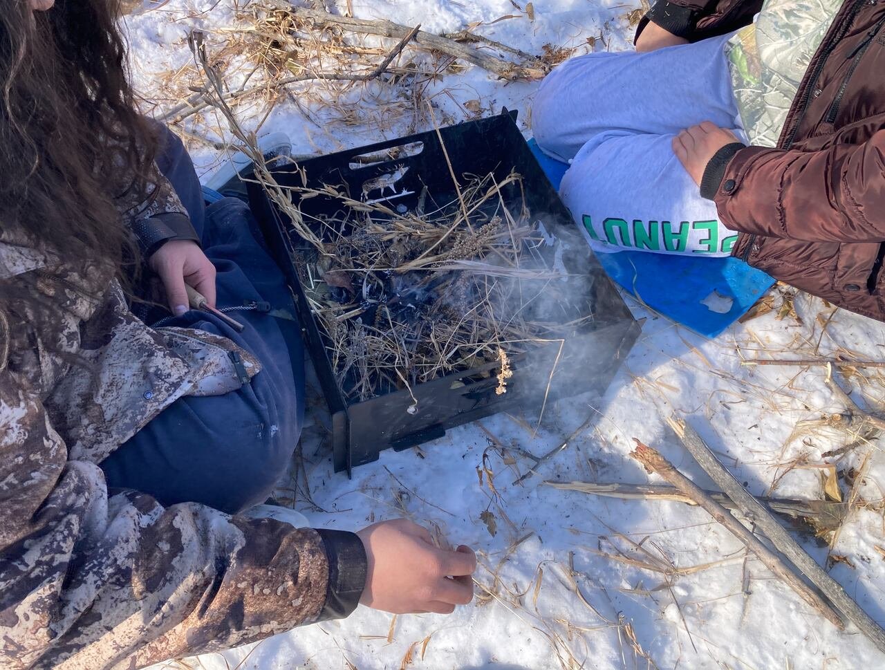 default-368 Young people kneeling on snowy ground around a burning metal box filled with dry grass and twigs.