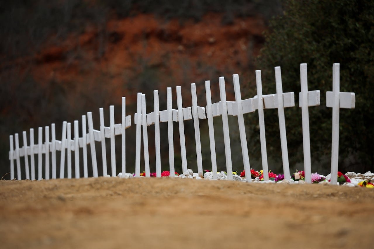 default-246 A row of 28 small white crosses in the dirt, a modest number of brightly coloured flowers at their base.