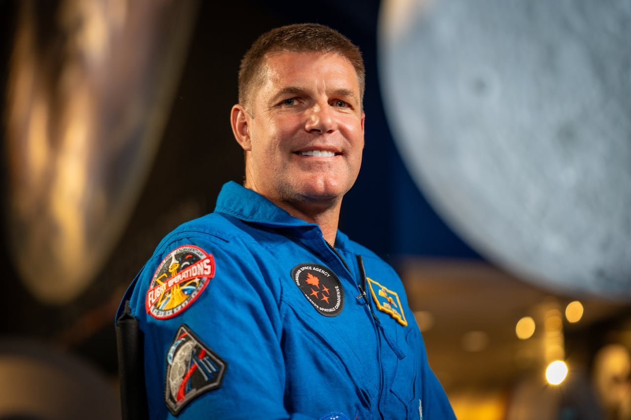 default-292 A man in a blue astronaut suit smiles with an image of the moon in the background.