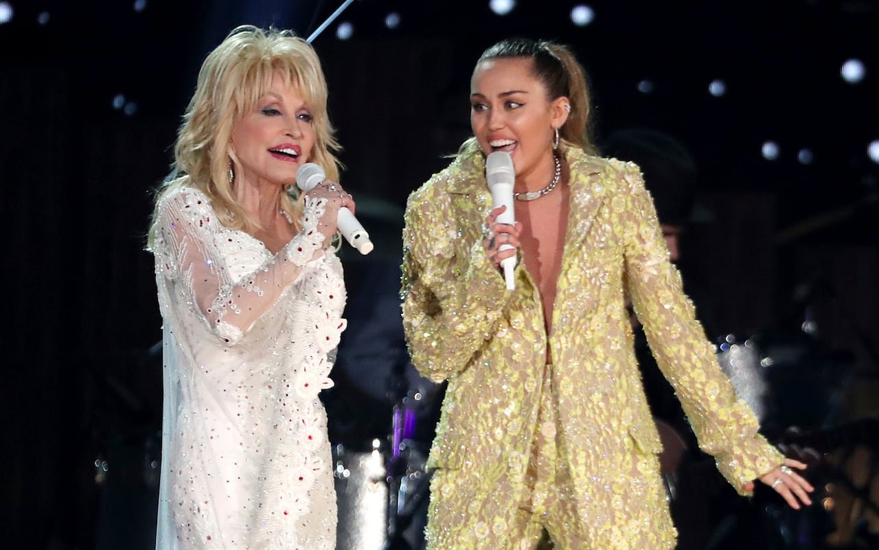 An elderly woman in a sparkling white outfit performs on stage with a younger woman in a shiny gold ensemble.