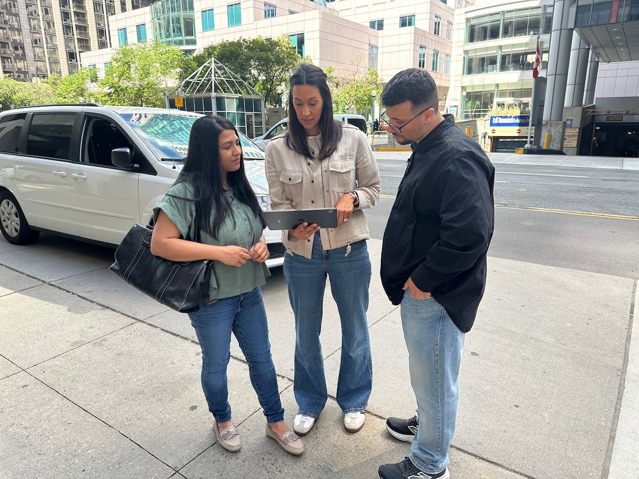 Three individuals reviewing car ads on a tablet with a parked van in the background.