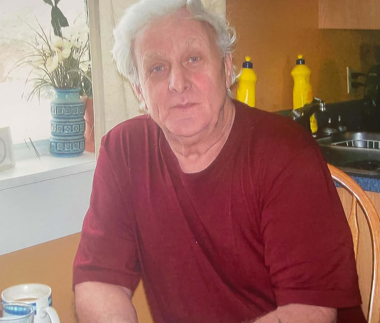 A man is sitting at a kitchen table. He's wearing a red t-shirt.