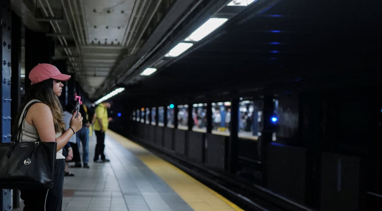 Woman stands on a dark subway platform holding a fan in front of her face